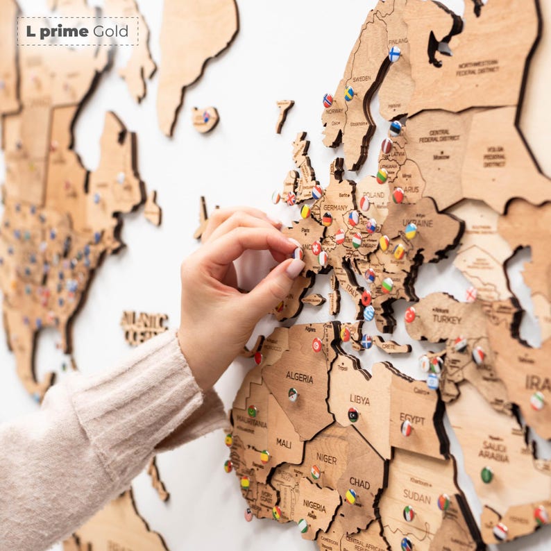 Pu&ograve; includere: Una mappa del mondo in legno con contorni di paesi e spilli. La mappa &egrave; realizzata in legno marrone chiaro ed &egrave; montata su una superficie bianca. Gli spilli sono colorati e rappresentano diversi paesi. Il testo "L prime Gold" &egrave; in alto a sinistra.