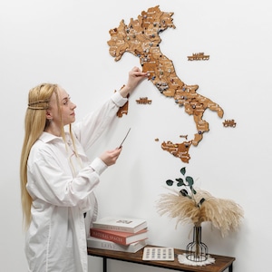 May include: A wooden map of Italy with pins marking locations, mounted on a white wall. The map is a light brown color. A person is pointing at the map with a pointer. Decorative items are on a wooden shelf below the map.