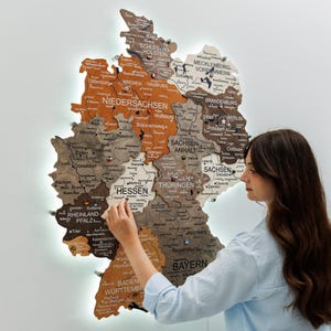 Peut inclure: Une grande carte murale en bois de l'Allemagne, composée de plusieurs tons, présentant des noms d'États, des villes et des frontières gravés au laser. La carte est fixée avec des entretoises, créant un effet de relief avec des nuances de bois.