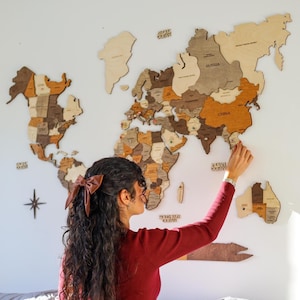 Peut inclure: Une grande carte du monde en bois, avec des pièces de pays individuelles dans différentes nuances de marron et de beige, est fixée sur un mur blanc. La carte comprend les noms des pays et des océans. Une personne place une épingle sur la carte.