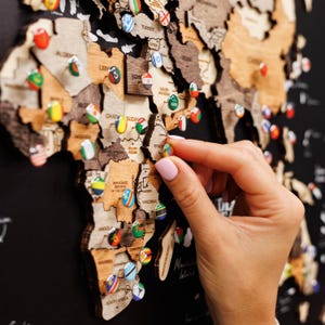 Peut inclure: Une carte du monde en bois avec des contours de pays et des épingles drapeaux colorées. Une main place une épingle sur le continent africain. La carte est fixée sur une surface noire, avec des noms de pays visibles.