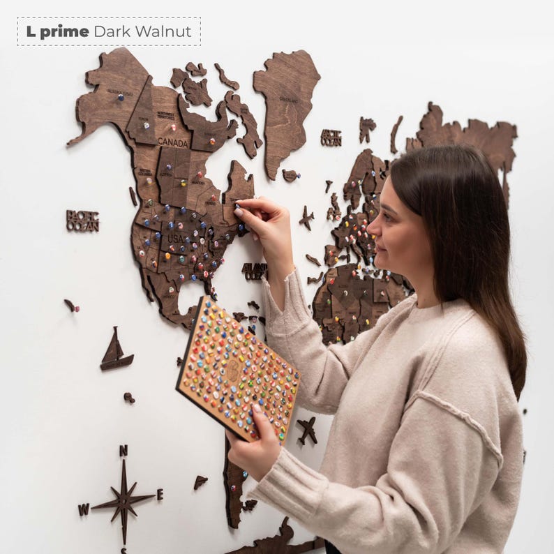 Pu&ograve; includere: Una mappa del mondo in legno di noce scuro montata su una parete bianca. La mappa &egrave; segmentata per paese e ha piccole puntine colorate inserite. Una persona sta inserendo una puntina sulla mappa. Una piccola bussola e una barca a vela in legno sono anche sul muro.