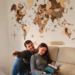 May include: A wooden world map wall decor with various shades of brown and tan is mounted on a white wall. A couple is sitting on a gray couch, with the woman reading a book. The man is holding a remote control.