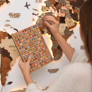May include: A person is placing a small flag pin on a wooden world map. The map is lit from behind, and the flags are arranged in a grid on a separate sheet of paper.