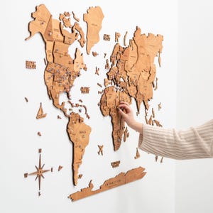 Puede incluir: Un mapa del mundo de madera con nombres de pa&iacute;ses y chinchetas, montado en una pared blanca. El mapa est&aacute; hecho de madera de color marr&oacute;n claro e incluye una rosa de los vientos y peque&ntilde;os recortes de barcos. Una mano coloca un alfiler en el mapa.