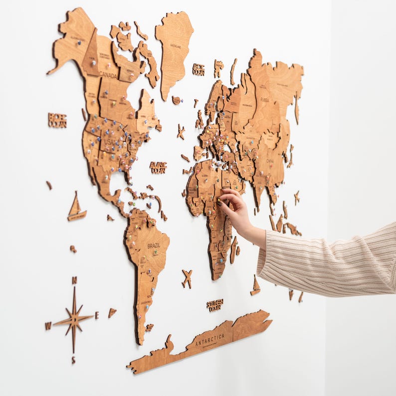 Puede incluir: Un mapa del mundo de madera con nombres de pa&iacute;ses y chinchetas, montado en una pared blanca. El mapa est&aacute; hecho de madera de color marr&oacute;n claro e incluye una rosa de los vientos y peque&ntilde;os recortes de barcos. Una mano coloca un alfiler en el mapa.