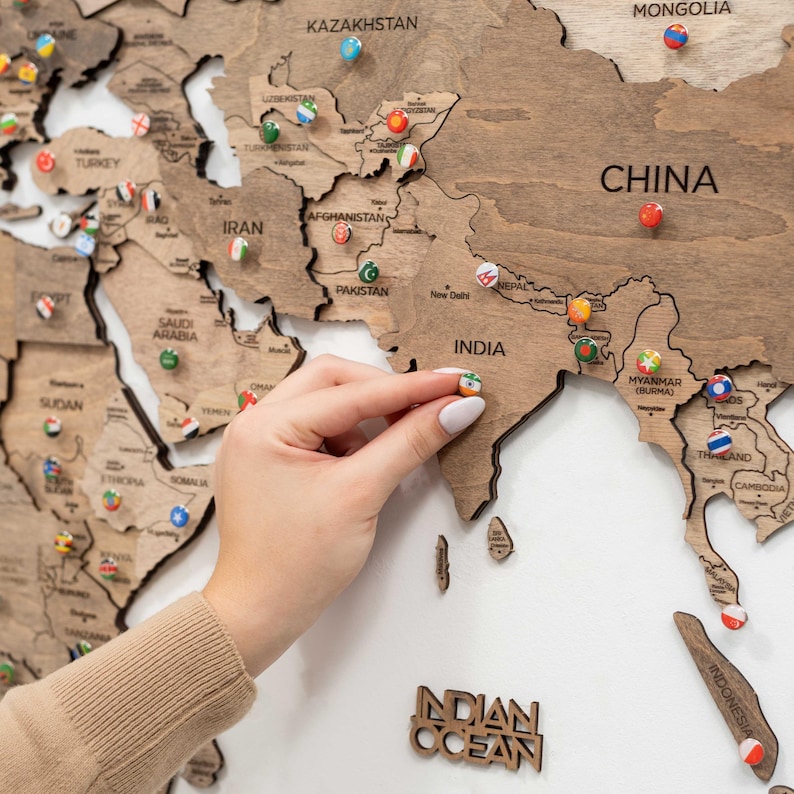 May include: A wooden world map wall art piece featuring laser-cut country borders and engraved labels for nations like China, India, and Kazakhstan. Small, round push pins decorated with various national flags are inserted into the wood surface.