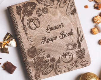 Puede incluir: Un libro de recetas rectangular de madera con un acabado marrón claro. La cubierta presenta ilustraciones grabadas con láser de varias verduras y el texto "Lauren's Recipe Book" en una fuente cursiva centrada en el frente.