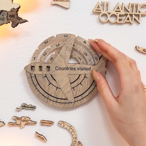 May include: A wooden travel tracker with the text "Countries Visited" and the number "17". The tracker is round with a compass design and a rotating dial. Other wooden map pieces are also visible.
