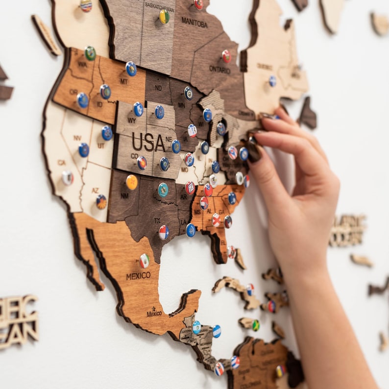 Pu&ograve; includere: Una mappa del mondo in legno con gli Stati Uniti evidenziati, con spilli con vari disegni di bandiere. La mappa &egrave; realizzata in diverse tonalit&agrave; di legno marrone ed &egrave; montata su una parete bianca. La parola "USA" &egrave; visibile.
