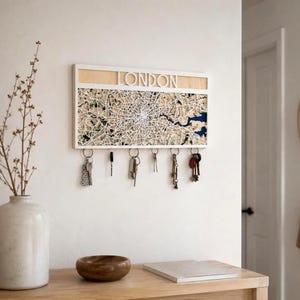 May include: A rectangular wall-mounted key holder featuring a white frame and a detailed map of London. The top section displays the word LONDON in white cut-out letters. Below, a map shows city streets and a blue river. Six metal hooks hold keys.
