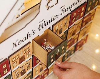 May include: A wooden advent calendar with small house-shaped drawers, each numbered. The calendar is painted in various colors, including red, green, and tan. The text "Noah's Winter Surprises" is written on the top. A hand is opening a drawer containing a small ornament.