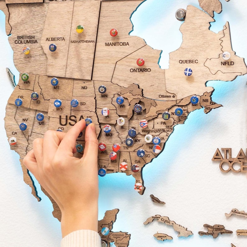 May include: A wooden world map with push pins marking different locations. The map is light brown with a blue outline. The map is mounted on a white wall. The map includes the United States, Canada, and parts of Mexico and Central America.
