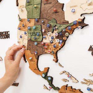 May include: A wooden wall map of North America featuring laser-cut borders and states. The map displays various shades of brown and green wood tones with small, circular pins placed across different regions, including the USA, Canada, and Mexico.