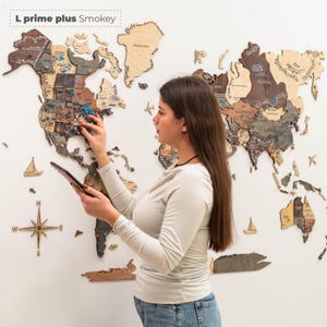 Pu&ograve; includere: Una mappa del mondo in legno dettagliata con contorni di paesi e variazioni di colore. La mappa &egrave; montata su una parete bianca, con una persona che tiene in mano un tablet e indica la mappa. La mappa include il testo "L prime plus Smokey".