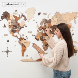 Puede incluir: Un mapa del mundo de madera en tonos marrones y beige, con chinchetas de colores, est&aacute; montado en una pared blanca. Una persona est&aacute; colocando chinchetas. El texto "L prime Multicolor" est&aacute; en la esquina superior izquierda.