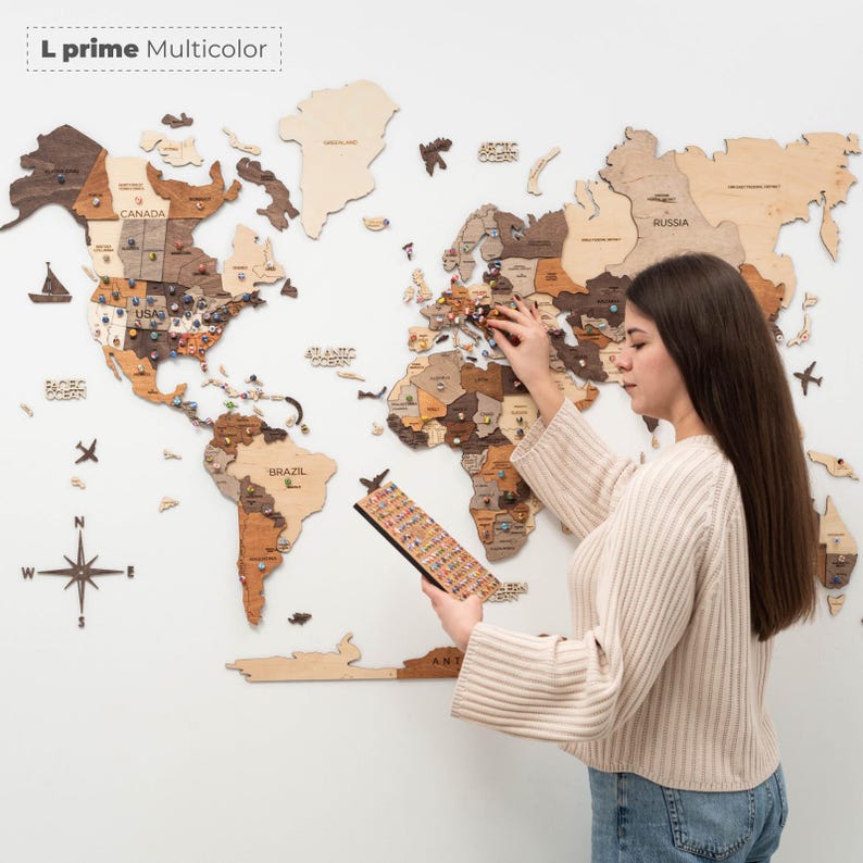 Puede incluir: Un mapa del mundo de madera en tonos marrones y beige, con chinchetas de colores, est&aacute; montado en una pared blanca. Una persona est&aacute; colocando chinchetas. El texto "L prime Multicolor" est&aacute; en la esquina superior izquierda.