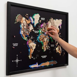 Op de afbeelding: Een houten wereldkaart met een frame en meerkleurige LED-verlichting die de continenten omlijnt. De kaart is gemonteerd op een zwarte achtergrond en bevat punaises. De kaart is gelabeld met de namen van de oceanen en kompasrichtingen.