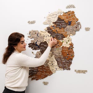 Op de afbeelding: Een houten kaart van Ierland in verschillende tinten bruin, beige en grijs, bevestigd aan een witte muur. De kaart is gelabeld met de namen van de provincies en heeft punaises. De woorden "Atlantic Ocean", "North Channel", "Irish Sea", "St. George's Channel" en "Celtic Sea" zijn ook zichtbaar.
