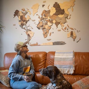 A cozy living room scene with a woman sitting on a brown leather sofa beside a large brown and white dog. Behind them, a wooden world map with country names in various wood tones is mounted on the wall, serving as a striking decor piece. The woman, dressed casually in jeans, boots, a Grand Canyon sweatshirt, and a cap, smiles while holding a mug. The space feels warm and inviting, perfect for relaxing and dreaming of travel adventures.