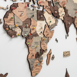 May include: A multi-layered wooden world map wall art featuring Africa and surrounding regions. The map consists of laser-cut wooden pieces in varying shades of brown and beige, with small circular pins displaying national flags placed on countries.