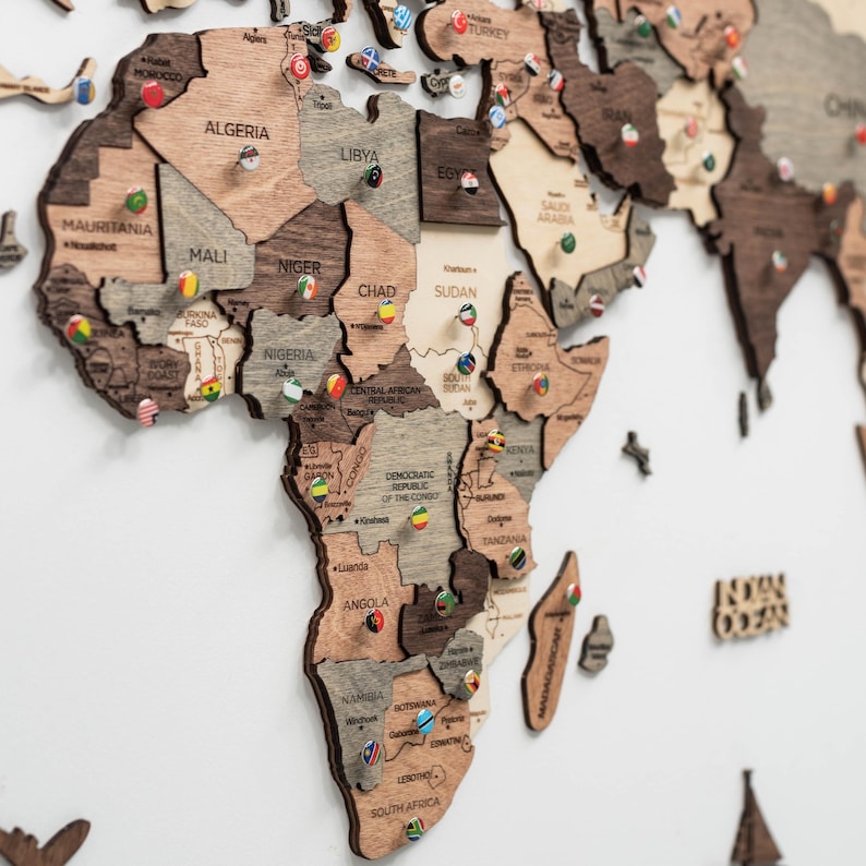 May include: A multi-layered wooden world map wall art featuring Africa and surrounding regions. The map consists of laser-cut wooden pieces in varying shades of brown and beige, with small circular pins displaying national flags placed on countries.