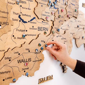 Könnte beinhalten: Eine Holzkarte der Schweiz mit detaillierten lasergeschnittenen Umrissen von Kantonen und Städten. Kleine, farbige Pins sind auf der Karte platziert und zeigen Standorte an. Das Wort "ITALIEN" ist unten sichtbar. Eine Hand platziert eine Stecknadel auf der Karte.