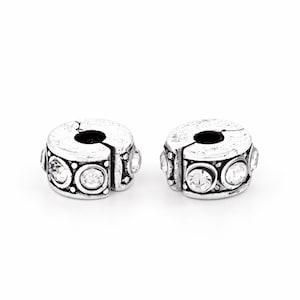 May include: Two silver-toned metal spacer beads with a hinged, openable design. Each bead features a textured, antiqued finish and is decorated with a series of clear, round, faceted crystals set in circular frames around the outer circumference.