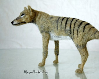 Puede incluir: Escultura de fieltro de un tilacino, también conocido como tigre de Tasmania, en posición de pie. El cuerpo es de color beige con rayas negras en la espalda y una larga cola. Firma del artista "Minzoo Needle Felting".
