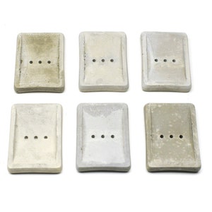 May include: Six rectangular concrete soap dishes in shades of grey are arranged on a white background. Each dish has three drainage holes. The soap dishes have a minimalist, modern design.