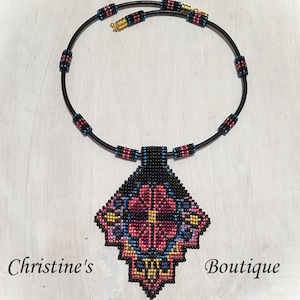 May include: A black beaded necklace with a large, colourful pendant featuring a geometric floral design. The pendant is made of black, red, pink, gold, and blue beads. The necklace is on a white background.