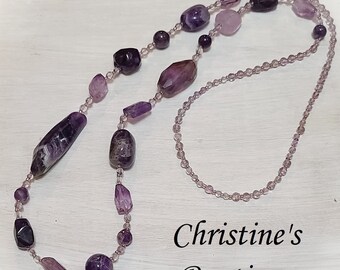 Könnte beinhalten: Eine lange violette Amethyst-Halskette mit Perlen in verschiedenen Größen und Formen. Die Halskette besteht aus kleinen, runden Perlen und größeren, unregelmäßig geformten Perlen. Die Halskette liegt auf einer weißen Oberfläche mit dem Text "Christine's Boutique" in Schwarz.