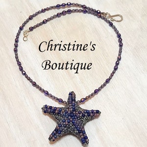 Puede incluir: Un collar de cuentas moradas y grises con un colgante de estrella de mar de cuentas. El collar está sobre una superficie de madera. El texto "Christine's Boutique" es visible en la imagen.