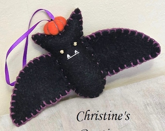 May include: A black felt bat ornament with purple stitching and a small orange pumpkin on its head. The bat has gold bead eyes and a white stitched mouth. A purple ribbon is attached for hanging. The text "Christine's Boutique" is at the bottom.
