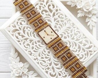 May include: A beaded bracelet in shades of brown, tan, and gold, featuring a square watch face design. The bracelet is approximately 2.5 cm wide and is displayed on a white, ornate tray with floral accents. The clasp is a matching beaded design.