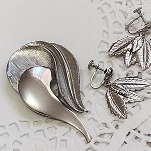 May include: A silver brooch in the shape of a stylized bird with a heart-shaped body and a long, curved tail. The brooch is paired with a set of silver earrings featuring a cluster of leaves.