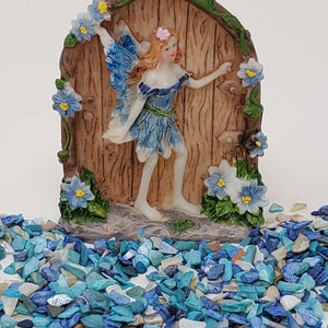 May include: A blue fairy figurine with wings, holding a flower, is standing in a doorway with a wooden door and blue flowers. The figurine is on a base with blue and white stones.