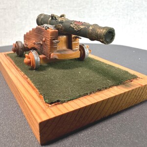 Peut inclure: Un modèle de canon miniature, avec un canon vert foncé et des détails dorés, monté sur un affût en bois marron et orange. Le modèle a de petites roues et repose sur une base en bois avec une surface en feutre vert.