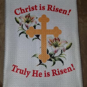 May include: White kitchen towel with a gold cross and white lilies printed on it. The text "Christ is Risen! Truly He is Risen!" is printed in red.