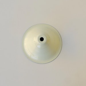 May include: A circular, off-white metal lamp shade with a smooth, glossy finish. The shade features a conical shape that tapers towards the centre, where a small, dark, circular opening is positioned for mounting purposes.