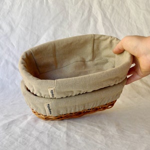 May include: Two beige oval-shaped wicker baskets with beige linen liners. The baskets are stacked on top of each other. The baskets have the word "mamieila" printed on the side.