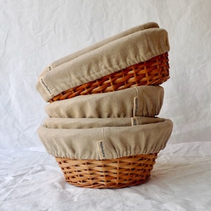 May include: Three beige linen-lined wicker bread baskets stacked on top of each other. The baskets are labeled "mainbäckerei" in black text.