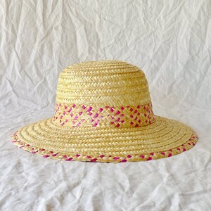 Puede incluir: Un sombrero de paja de ala ancha con una banda tejida rosa alrededor de la corona.