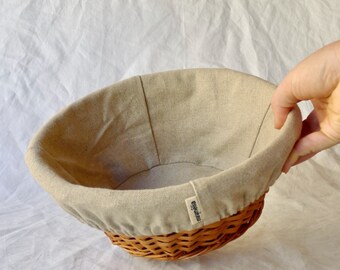 May include: A round, woven wicker bread basket with a beige linen liner. The liner has a small, blue label that says "Eugénie" on it.