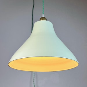 Könnte beinhalten: Eine glockenförmige Pendelleuchte mit einer matten, blass-minzgrünen Außenseite und einer warmgelben Innenseite. Die Leuchte hat eine kleine messingfarbene Metallkappe an der Oberseite und hängt an einem dünnen, gedrehten grünen Textilkabel.