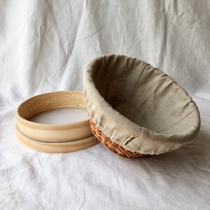 May include: Two round, woven baskets. One is a brown wicker basket with a beige linen liner. The other is a wooden basket with a woven top. The baskets are on a white fabric background.