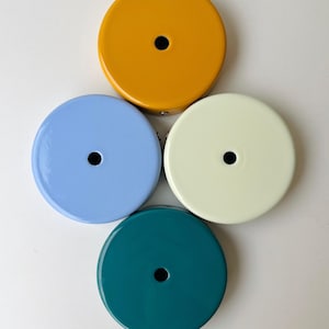 Puede incluir: Cuatro cubiertas de techo redondas y coloridas, vista superior. Los colores son amarillo mostaza, azul claro, crema y verde azulado. Cada una tiene un agujero negro centrado. Estas cubiertas decorativas son para luces de techo.