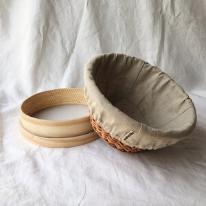May include: A round, wooden proofing basket with a removable liner. The basket is made of woven wicker and has a light brown color. The liner is made of a light beige fabric and has a drawstring closure.