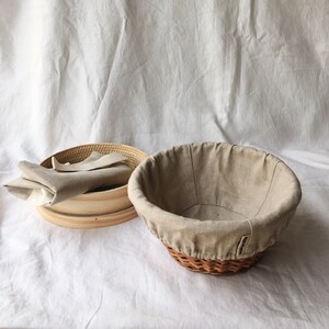 May include: A set of two beige linen-lined bread baskets. One basket is round and woven, the other is a wooden round with a removable linen liner.