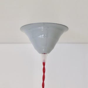 Pode incluir: Uma canopla de teto cinza-claro em forma de sino com acabamento liso e brilhante. Um cabo elétrico de tecido trançado vermelho vibrante estende-se para baixo a partir do centro, fixado por um pequeno componente de alívio de tensão de plástico.